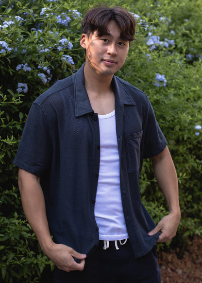 Man wearing white ribbed tank top and navy shirt, standing outdoors by green bushes with blue flowers.
