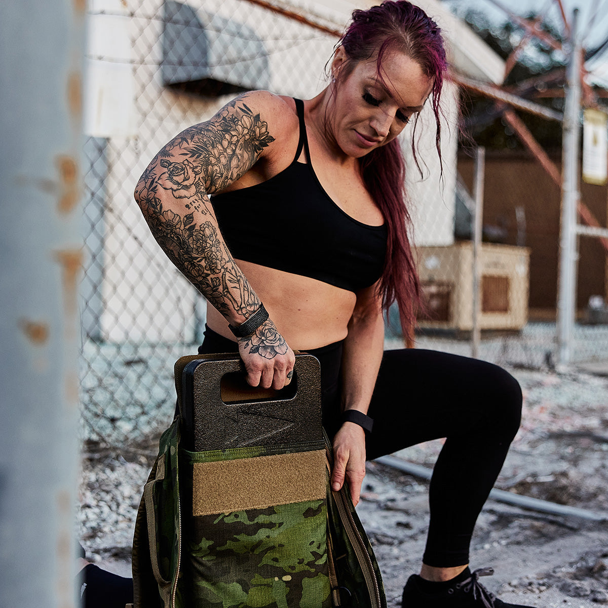 Tattooed woman in athletic wear loading a ruck plate into a camo workout backpack outdoors