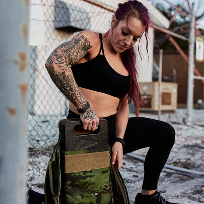 Tattooed woman in athletic wear loading a ruck plate into a camo workout backpack outdoors