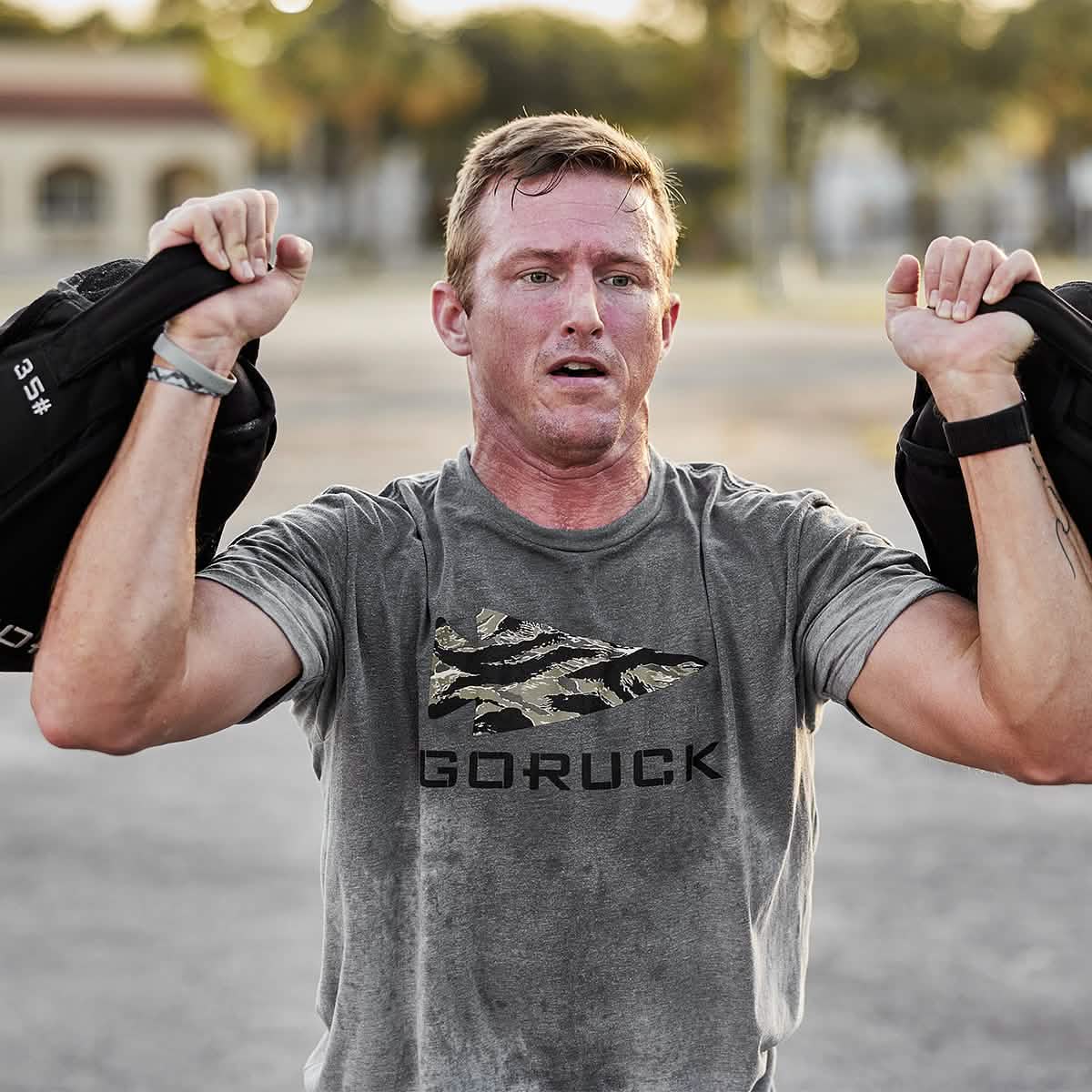 Athletic man lifting weights outdoors in a camo GORUCK T-shirt, men's fitness lifestyle