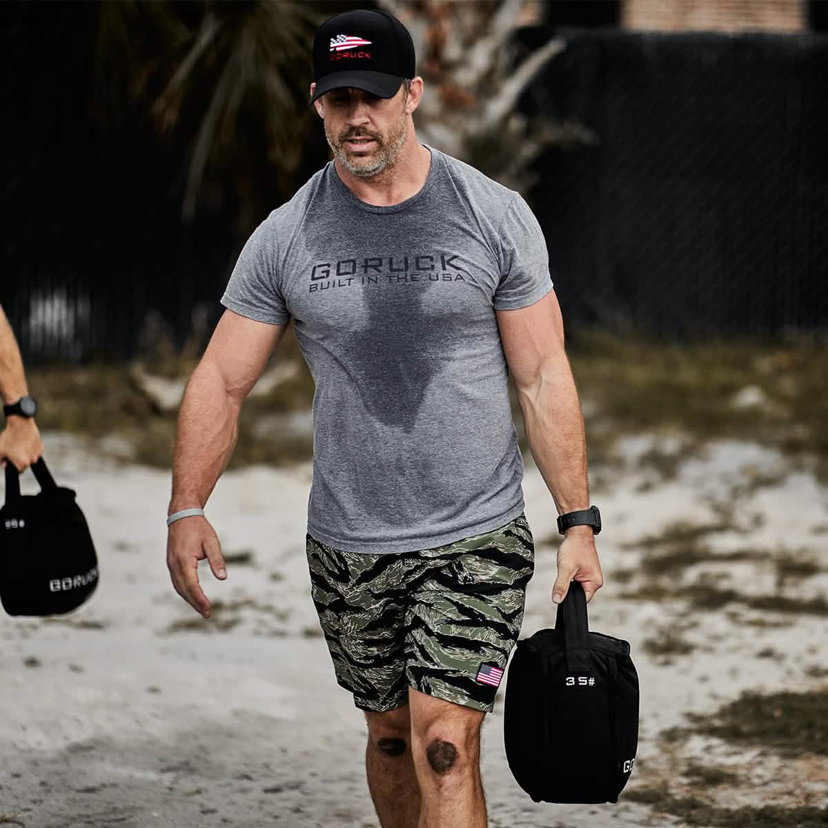 Athletic man in GORUCK t-shirt and camo shorts carrying weighted bag outdoors, Mann marketplace style