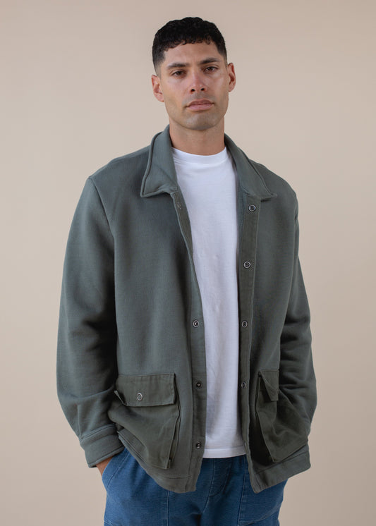 Man modeling a green worker's jacket over a white t-shirt and blue pants, minimalist background